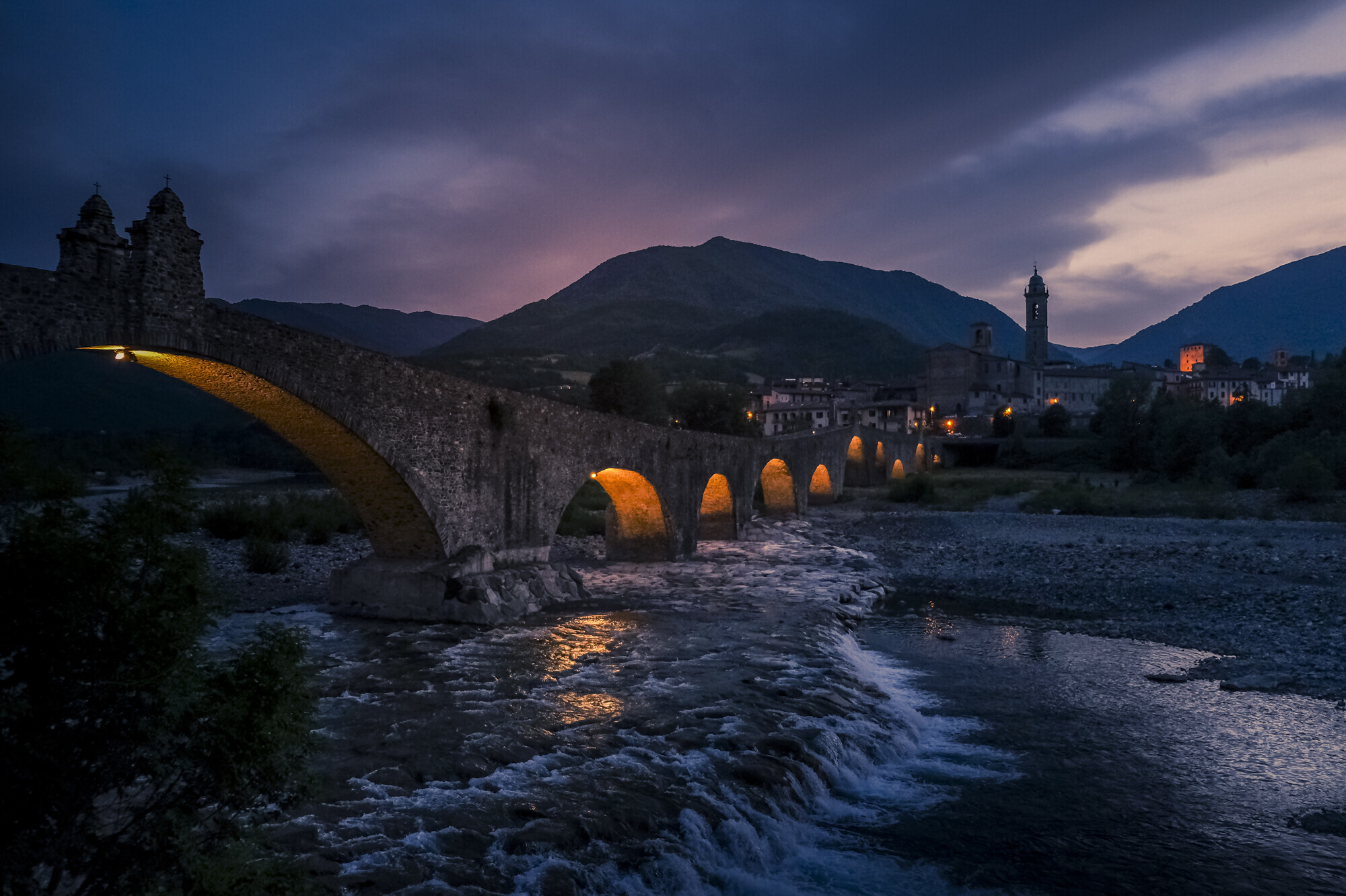 31 Sera a Bobbio QUARTA CLASS MICHELANGELO BIAGINI
IL BACCHINO
Nella sera brillano le luci sul Ponte Gobbo e sulle schiumose onde della Trebbia. In lontananza il paese con lo svettante campanile. Tutto  silente,
solo il fiume dialoga con il ponte. Un mor